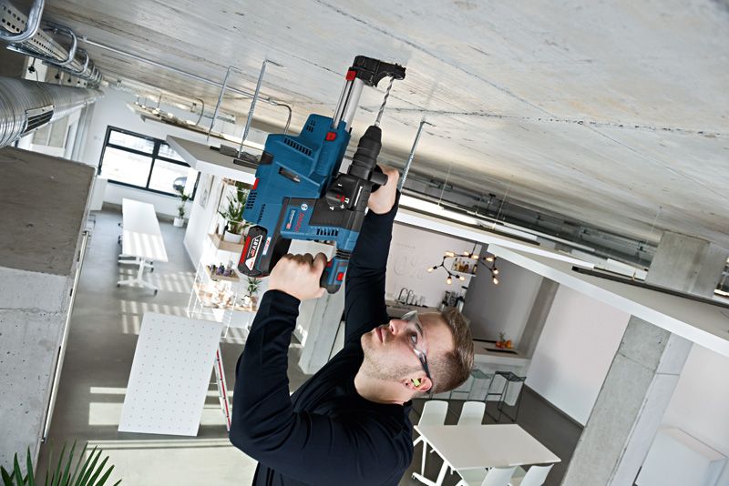 GBH 18V-26F A person wearing safety equipment drills into a concrete ceiling in a modern workspace.