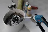 Electric mixer blending paint in a bucket beside assorted paint bottles and a roller.