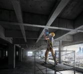 A worker wearing safety equipment drills into a concrete ceiling on scaffolding.