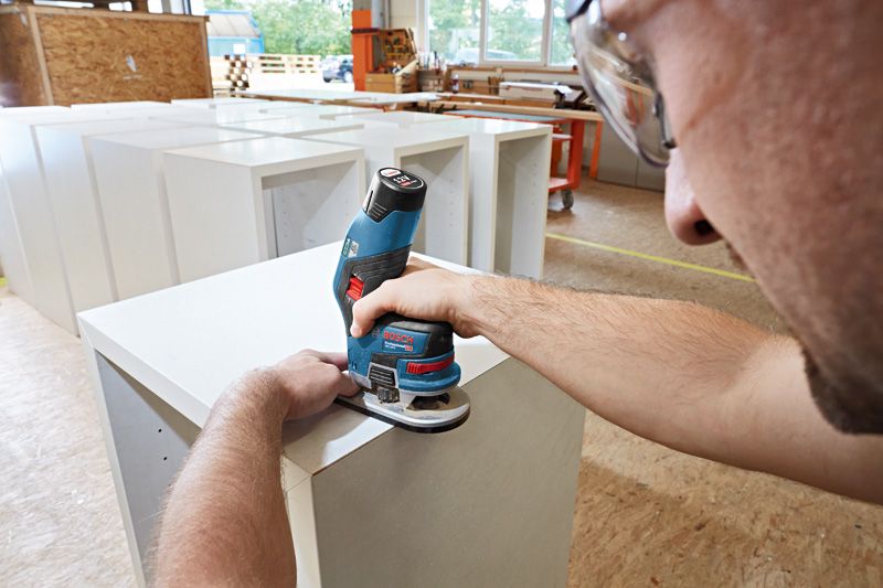 A person wearing safety equipment shapes wood edges using a cordless palm router.