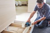 A person uses a cordless planer to smooth a wooden board on a jobsite floor.