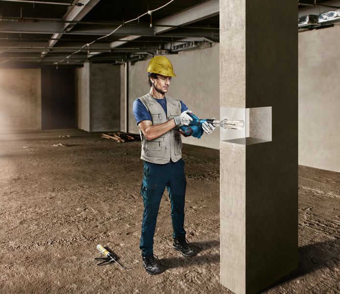 A person wearing safety equipment drills into a concrete pillar in a construction area.