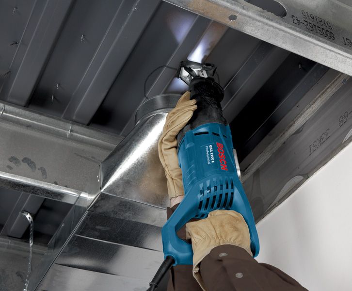 A person wearing safety equipment uses a reciprocating saw to cut sheet metal overhead.