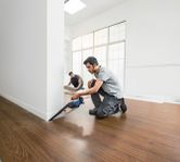 A person cuts flooring near a wall with a cordless saw while another installs floor panels.