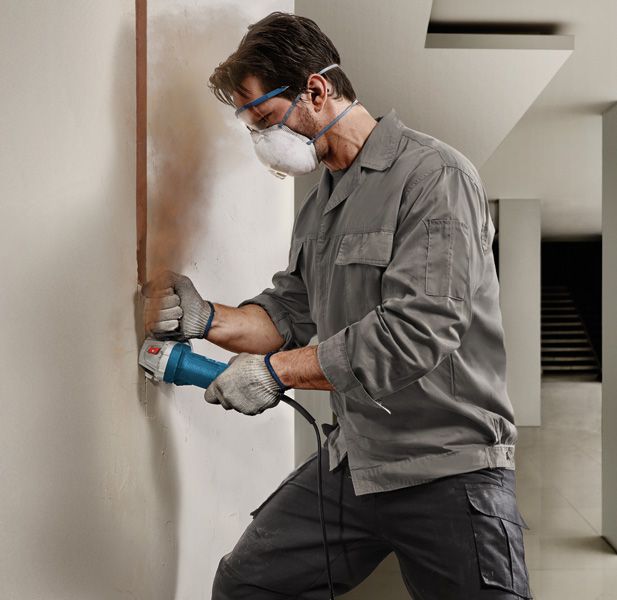A person wearing safety equipment uses a power tool to cut a vertical groove in a wall.