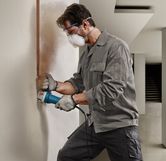 A person wearing safety equipment uses a power tool to cut a vertical groove in a wall.