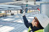 A person wearing safety equipment uses a cordless impact wrench on a metal beam.