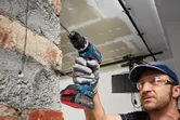 A person wearing safety equipment drills into a brick wall with a cordless power drill.