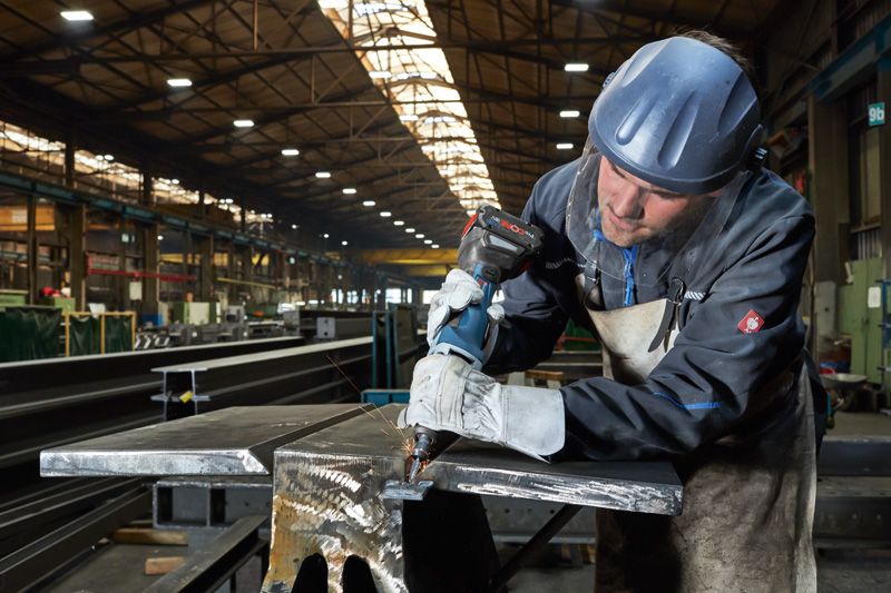 GGS 18V-23 PLC A person wearing safety equipment drills into a metal beam in a factory.