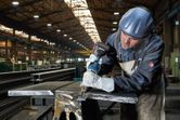 A person wearing safety equipment drills into a metal beam in a factory.