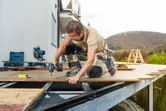 A person wearing safety equipment uses a cordless impact driver to fasten decking boards.