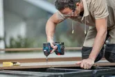 Person wearing safety equipment uses a cordless impact driver to fasten a screw into wood.