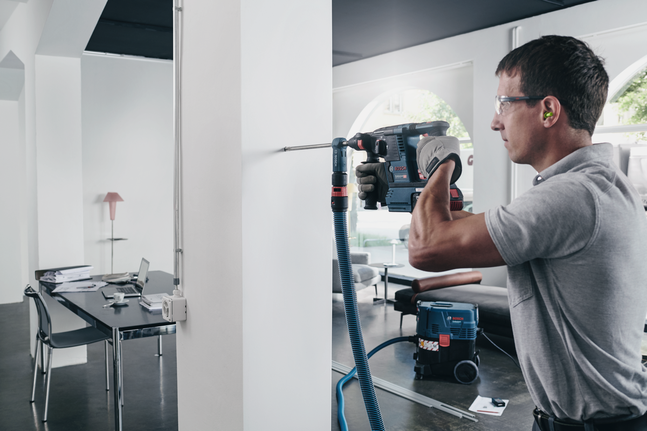SDS plus-9 Speed Clean A worker wearing safety equipment drills into a white wall in a modern office.