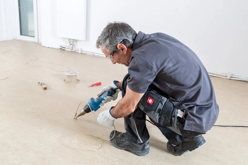 A person wearing safety equipment uses a heat gun to work on flooring.