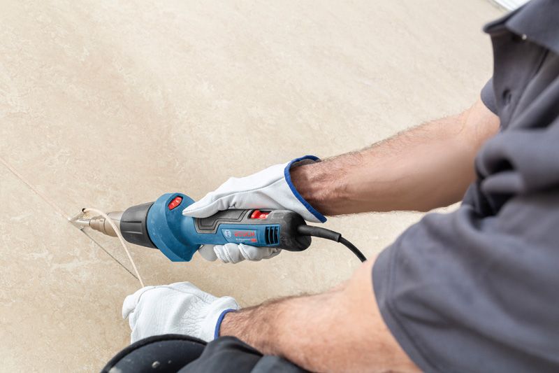 A person wearing safety equipment uses a heat gun to strip material from a surface.