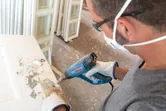 A person wearing safety equipment uses a heat gun to strip paint from a door.
