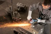 A person wearing safety equipment grinds a large metal component with an angle grinder.