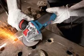 A person wearing safety equipment uses an angle grinder to cut through metal, creating sparks.