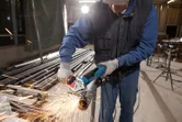 A person wearing safety equipment grinds a metal pipe with an angle grinder, producing sparks.