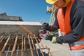 A person wearing safety equipment uses an angle grinder to cut rusted rebar on a construction site.