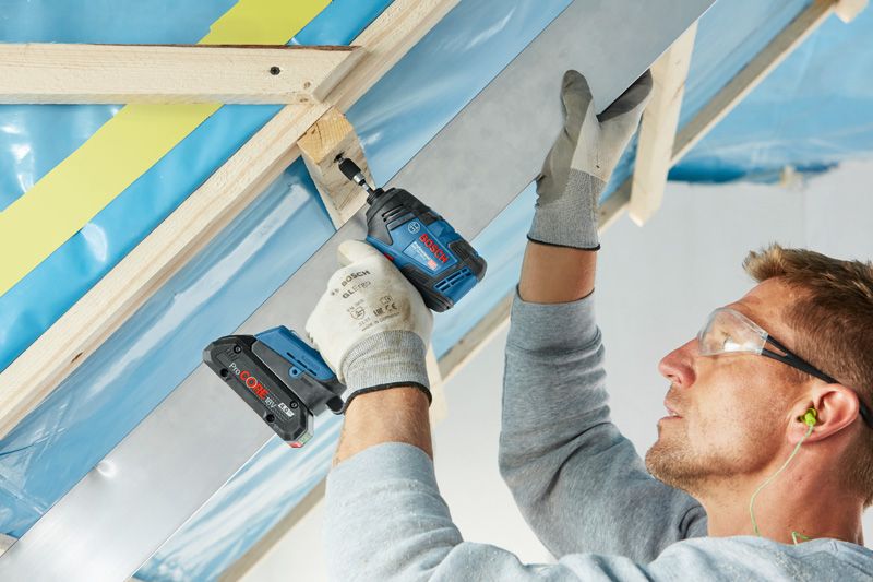 A person wearing safety equipment fastens a metal beam to a wooden frame with a power drill.