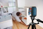 A person installs a cabinet in a kitchen using a laser leveling tool on a tripod.
