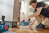 A person wearing safety equipment uses a cordless vacuum cleaner to clean wood shavings.
