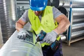 A person wearing safety equipment fastens a vent cover to a metal duct with a power tool.