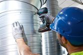 A person wearing safety equipment uses a cordless impact driver on a metal air duct.