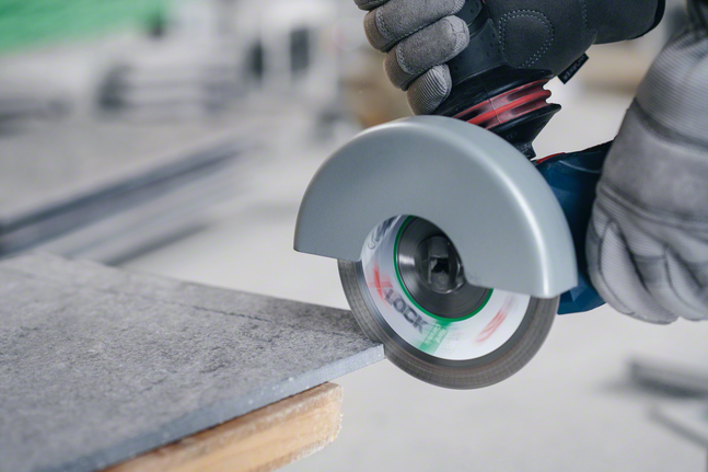 X-LOCK Standard for Ceramic diamond cutting disc 125x22.23x1.6x7 A person wearing safety equipment cuts a tile with an angle grinder.