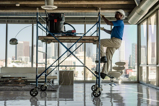 Person wearing safety equipment climbs a scaffold in a modern office with power tools above.