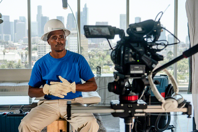 Person wearing safety equipment sits in front of a video camera with city buildings in the background.