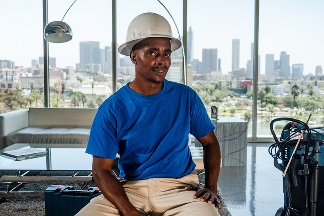 Person wearing safety equipment sits indoors beside power tools with a city skyline in the background.