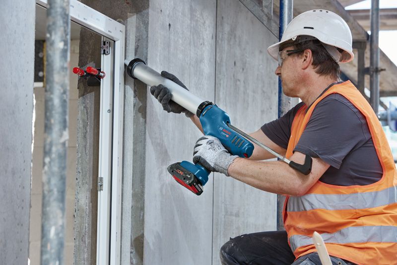 GCG 18V-600 A person wearing safety equipment applies sealant to a window frame using a cordless caulk gun.