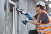 A person wearing safety equipment applies sealant to a window frame using a cordless caulk gun.