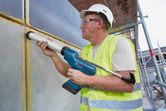 A person wearing safety equipment applies sealant to a wall joint with a cordless caulk gun.