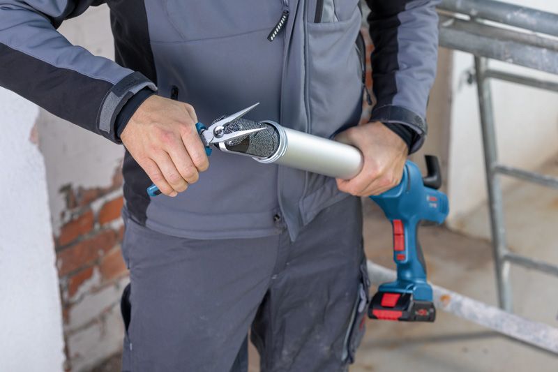 A person wearing safety equipment cuts the tip off a caulk tube for a cordless caulk gun.