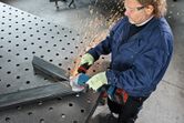 A worker wearing safety equipment grinds metal beams on a perforated work table.