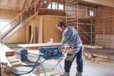 A person operates a table saw to cut wood in a partially constructed building.