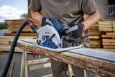Person wearing safety equipment cuts a rough wooden plank with a circular saw.