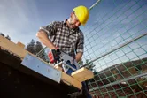 A person wearing safety equipment uses a cordless circular saw to cut wood on a construction site.