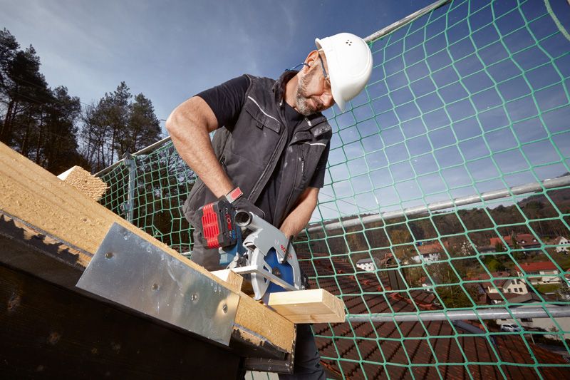 A person wearing safety equipment uses a circular saw to cut wood on a rooftop.