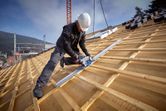 A person wearing safety equipment cuts wood on a sloped roof using a track saw.