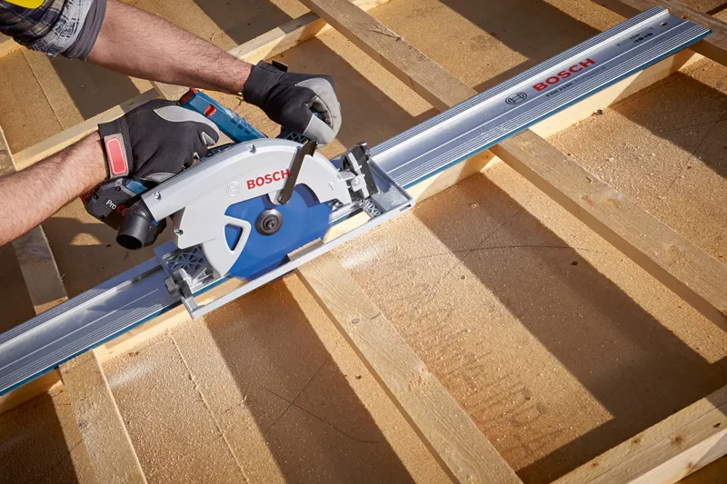 A person wearing safety equipment uses a cordless circular saw to cut wooden boards.