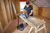 A person wearing safety equipment cuts chipboard with a cordless circular saw on a guide rail.