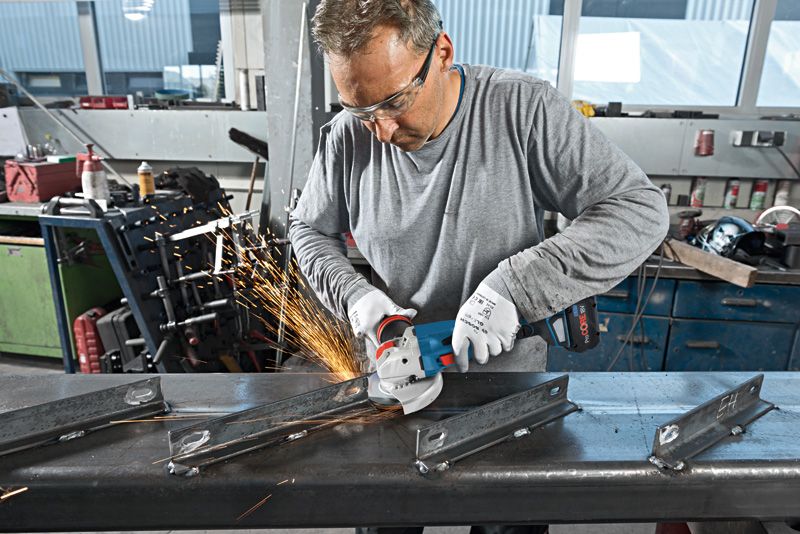 GWX 18V-15 C A worker wearing safety equipment grinds metal beams using a cordless angle grinder.