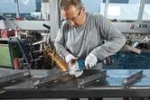 A worker wearing safety equipment grinds metal beams using a cordless angle grinder.