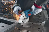 A person wearing safety equipment grinds a metal beam with a cordless angle grinder.