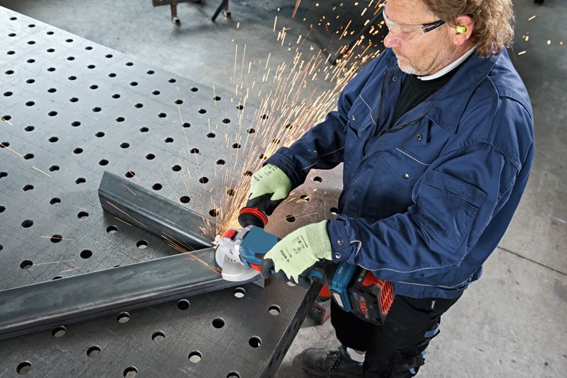 A person wearing safety equipment grinds metal tubing, sparks flying from a cordless angle grinder.