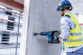 A person wearing safety equipment uses a cordless rotary hammer to drill into concrete.